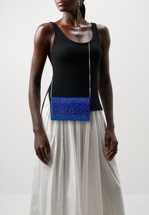 Kvinna som bär en svart ärmlös topp och vit kjol, och bär en liten blå pärlbead crossbody-väska med en silverkedjesnor.