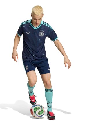 Tânărul jucător de fotbal bărbat, în echipamentul Germaniei în nuanțe de bleumarin și verde-teal, controlează o minge multicoloră cu piciorul drept pe un fundal alb.