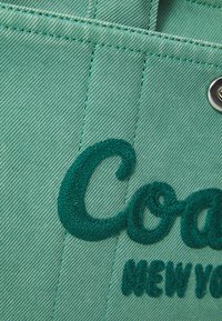 Šviesiai žalias džinsinis audinys su kontrastinga tamsiai žalia siuvinėta "Coach" ir "New York" užrašu. Matomas siuvimo ir inkarų detalės.