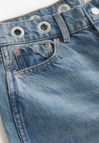 Shorts din denim de culoare albastră deschisă, cu nituri din cupru, grommeturi din argint și cusături contrastante de culoare portocalie. Buzunarul din față este vizibil.