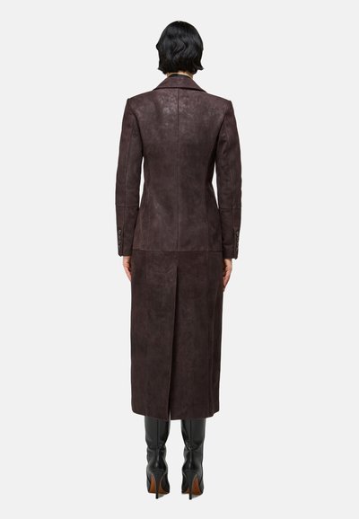 Femme aux cheveux courts noirs portant un long manteau en cuir marron foncé et des bottes à talons noires, vue de dos sur un fond blanc.