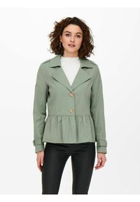 Giacca peplum verde chiaro con colletto a punte, due bottoni prominenti, maniche lunghe e una texture liscia. Abbinata a pantaloni neri.