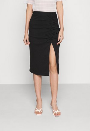 Jupe midi noire avec texture froncée, fente sur le côté et silhouette ajustée, assortie à des sandales blanches à lanières.