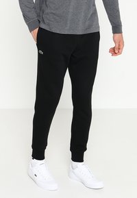 Joggers de algodón negros con puños elásticos, que presentan un logotipo sutil en el muslo. Combinados con zapatillas blancas, que complementan una camiseta de manga larga gris.