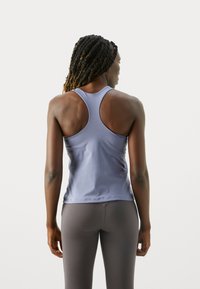 Kvinde med fletninger iført en lyseblå racerback tanktop og grå leggings, stående mod en ensfarvet hvid baggrund.