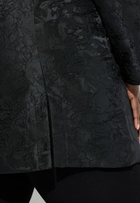 Sort tekstureret stof med blomsterbroderimønster, med skræddersyet snit og sideslids for nem bevægelse.