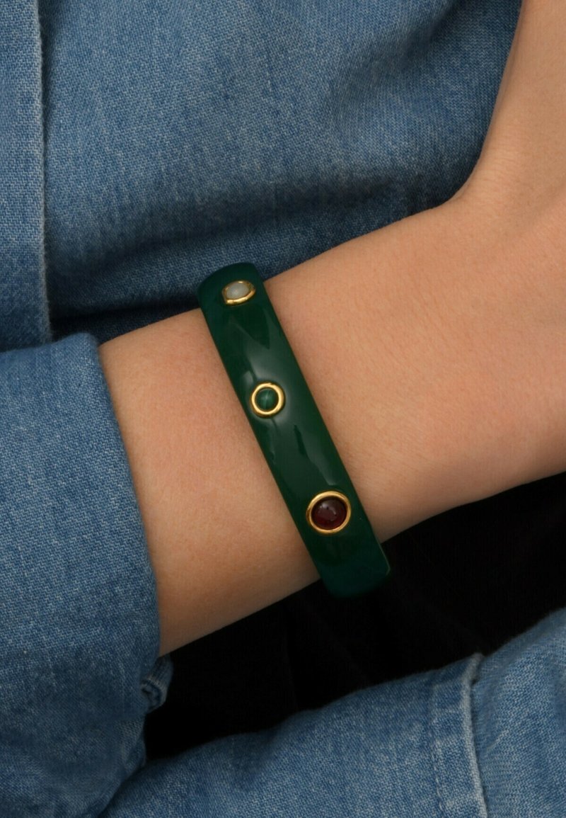 Bracelet en résine verte avec trois clous dorés : un vert, un rouge et un transparent. Porté au poignet, associé à un tissu en denim.