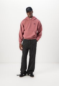 Hoodie rose surdimensionné avec poche et logo noir, associé à un pantalon noir texturé avec fermeture éclair et détails en frange. Casquette noire sur la tête.