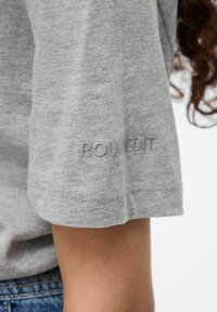 Manica di una maglietta in cotone grigia con testo ricamato discreto, indossata da una persona con capelli ricci e jeans in denim blu.