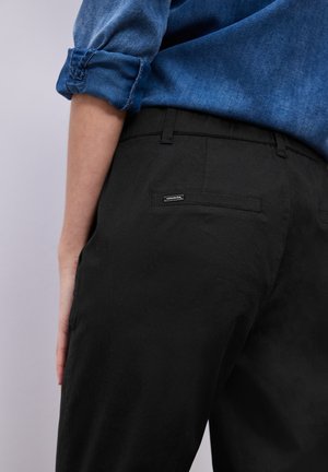 Personne portant une chemise en denim retroussée et un pantalon noir avec une poche passepoilée au dos et une petite étiquette métallique sur la poche.