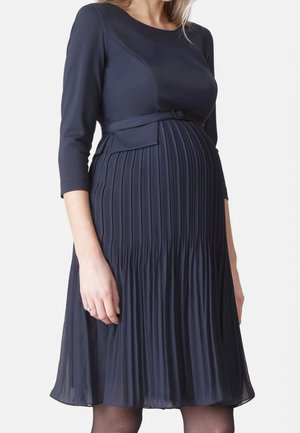 Zwangere vrouw die een marineblauwe jurk tot op de knie draagt met driekwart mouwen en een geplooide rok, haar zwangerschapsbuik tonend.