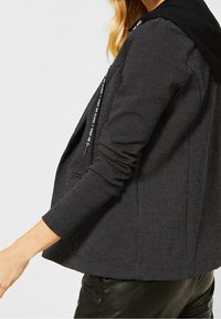 Veste noire avec un motif texturé en points, dotée d'une capuche noire et d'une fermeture éclair avec un logo blanc sur le côté. Manches et ourlet ajustés.