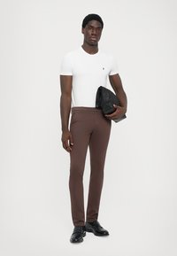 Biela priliehavá tričko, hnedé slim-fit nohavice, čierne kožené topánky a čierna kožená bunda držaná v jednej ruke. Jednoduché pozadie.