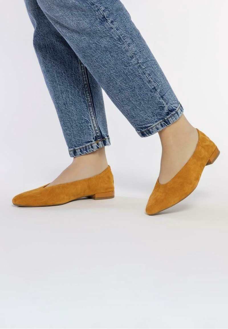 Pieds portant des chaussures plates en daim orange associées à un jean bleu en denim sur fond blanc uni.