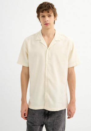 Camisa de botones beige de manga corta con cuello, que presenta una tela texturizada y dobladillo recto, combinada con jeans gris oscuro.
