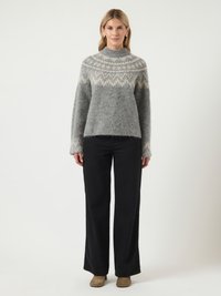 Vrouw in een grijs gemusterde gebreide trui, zwarte wijde broek en bruine suède enkellaarsjes, staande tegen een witte achtergrond.
