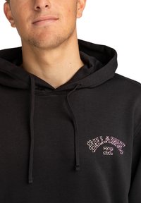 Sudadera negra hecha de un tejido suave y transpirable. Cuenta con una capucha con cordón y un logotipo frontal con patrones en rosa y blanco. Diseño sencillo y casual.