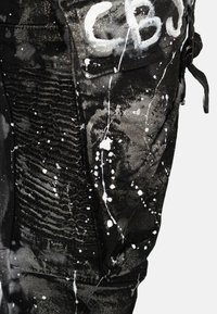 Pantalons noirs avec une texture, ornés d'un design de peinture éclaboussée en blanc, et dotés d'un élastique sur le côté pour un look ajusté.