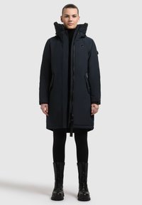 Marineblå parka med en hætte foret med faux pels, lynlås foran, sidelommer og ribbet detaljering. Sammen med sorte leggings og kraftige sorte støvler.