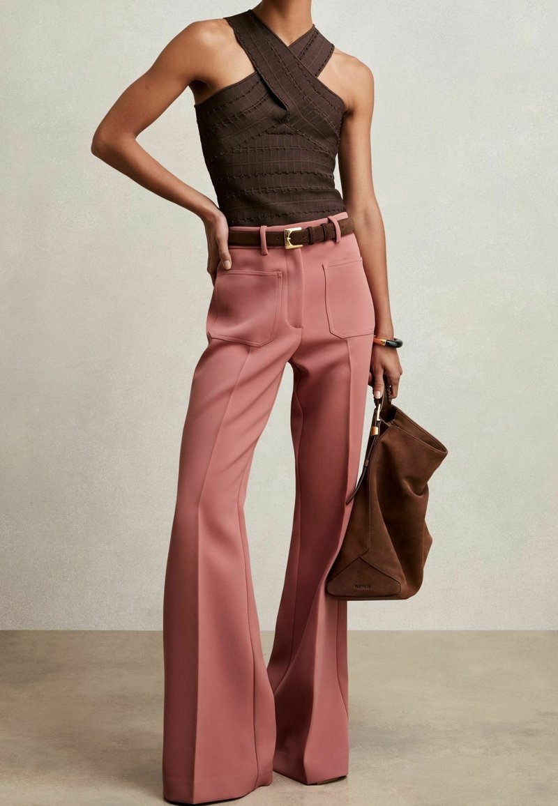 Femme portant un haut sans manches marron foncé avec des bretelles croisées, un pantalon large rose taille haute, tenant un sac à main marron décontracté dans une main.