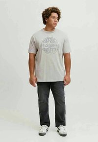 T-shirt de algodão cinza claro com uma impressão de texto circular que diz "Roupas de Qualidade Superior." Combinado com jeans cinza escuro e ténis brancos.