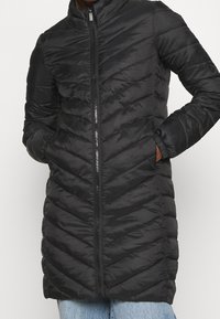 Cappotto puffer isolato nero con design a trapunta a zigzag, colletto alto, zip frontale e tasche laterali. Finitura in tessuto liscio e lucido.
