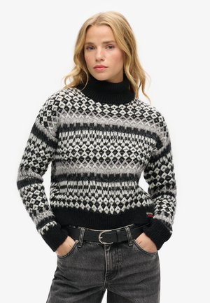 Junge Frau mit blondem Haar, die einen schwarz-weiß-grau gemusterten Strickpullover und dunkelgraue Jeans trägt und die Hände in den Taschen hat.