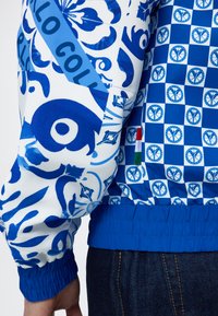 Μανίκι και πλάτη μπλε και λευκοντυμένου μπουφάν με καρό σχέδιο ειρηνικού συμβόλου και ελαστική μπλε μανσέτα, φορεμένο με σκούρα τζιν.