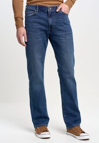 Vaqueros de mezclilla azul para hombre con corte recto, diseño clásico de cinco bolsillos, ligeras arrugas en la tela y cierre con botón, combinados con zapatillas deportivas marrones.