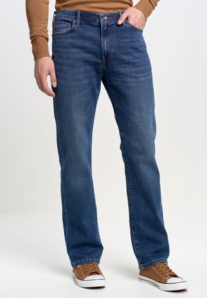 Jeans in denim blu da uomo con taglio dritto, design classico a cinque tasche, leggeri segni di usura e chiusura con bottoni, abbinati a sneakers marroni.