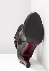 Botte en cuir noire à talon moyen avec fermeture éclair latérale, semelle inclinée vers le haut, reposant contre un mur blanc et une surface.