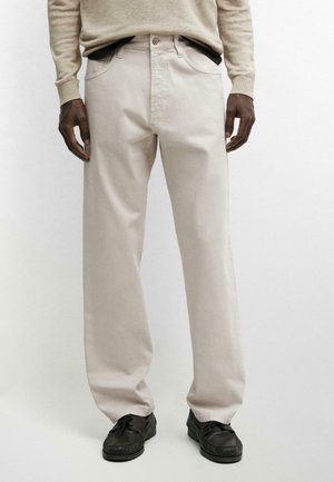 Mann trägt beige gerade geschnittene Hose, beiges langärmeliges Oberteil und schwarze Schnürschuhe, steht vor weißem Hintergrund.