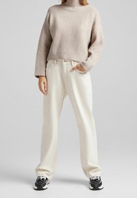 Lichtbeige gebreide coltrui met geribde manchetten, gecombineerd met loszittende witte jeans en zwarte sneakers met witte accenten.