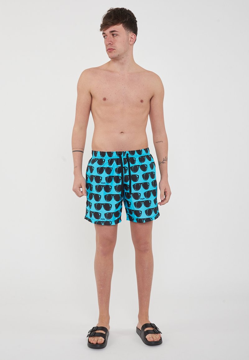 Costumi da bagno in tessuto azzurro chiaro con un motivo ripetuto di occhiali da sole neri. Comoda cintura elastica con cordino. Sandali neri indossati.