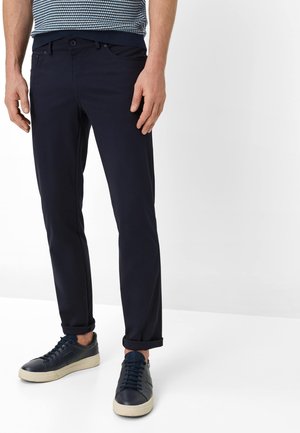 Mann trägt dunkelblaue Slim-Fit-Hosen, an den Bündchen hochgekrempelt, kombiniert mit schwarzen Sneakern mit weißen Sohlen und einem gestreiften Hemd.