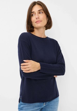 Vrouw met kort bruin haar, draagt gouden ringoorbellen, een marineblauwe langemouwen sweater en lichtblauwe jeans. Ze staat met gekruiste armen en kijkt naar rechts.