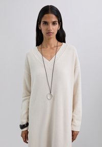 Vestido largo de manga larga color crema con escote en V, presenta puños ribeteados y un pespunte decorativo a lo largo del escote. Correa delgada marrón con un colgante circular.