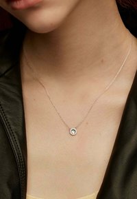 Collar de cadena de plata con un pendant circular, que presenta una gema clara rodeada de un halo de piedras preciosas. Lleva puesto contra una blusa de cuero oscura.