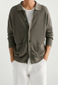 Olive-green ribbed cardigan with a classic collar, front button closure, and two front pockets, paired with white trousers.