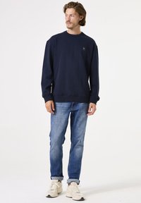 Sudadera azul marino con cuello redondo y puños acanalados, combinada con jeans azul claro y zapatillas beige. Branding mínimo en el pecho.
