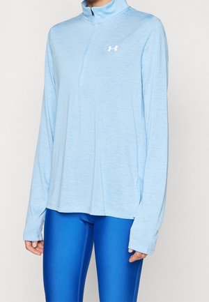 Jasnoniebieska sportowa bluza z długim rękawem, krótkim zamkiem i logo Under Armour, noszona z jasnoniebieskimi legginsami na osobie.