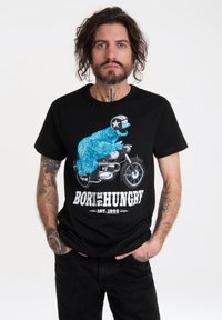 Camiseta de algodón negra con un personaje de dibujos animados azul en una motocicleta, con el texto "NACIDO PARA TENER HAMBRE" y "EST. 1966" en blanco.