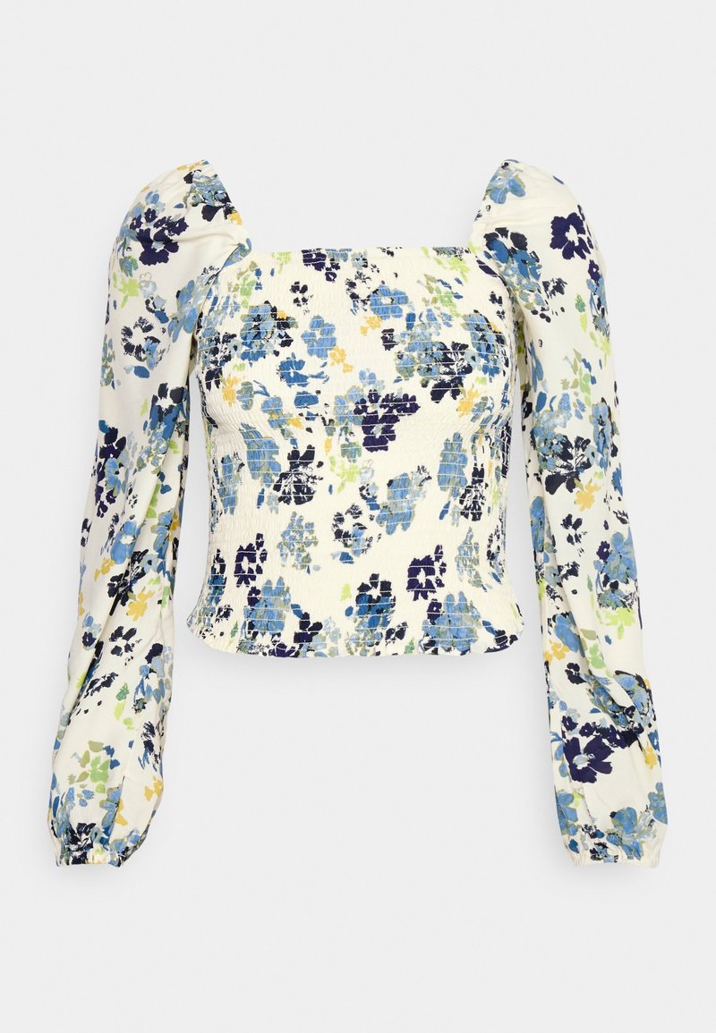 Haut blanc à manches longues smocké avec imprimé floral bleu, vert et jaune et épaules bouffantes sur fond uni.