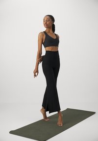 Black sports bra and high-waisted flared black leggings on a model, standing barefoot on a dark green yoga mat with a neutral background.