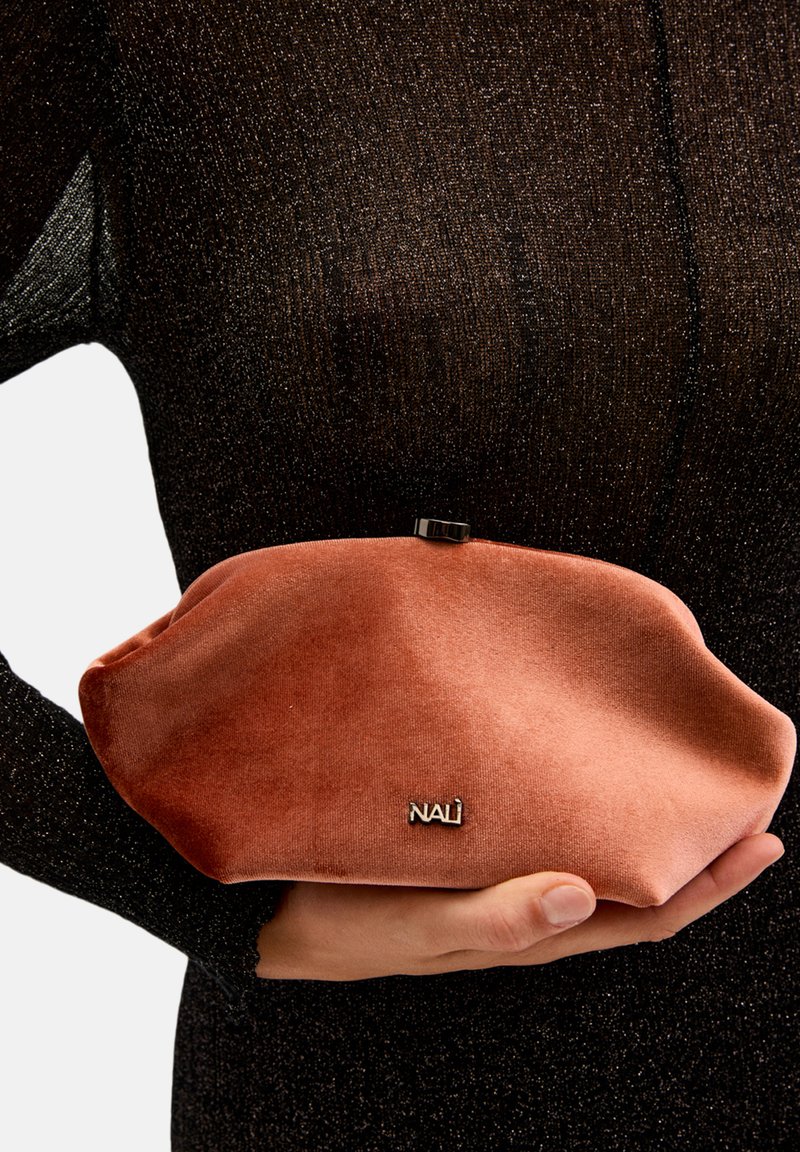 Rosa sammetclutch med rund form, metallfäste och präglad logotypdetalj. Mot en svart strukturerad bakgrund.