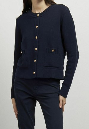 Femme portant un cardigan bleu marine boutonné avec des boutons dorés et un pantalon bleu marine assorti, debout devant un fond clair uni.