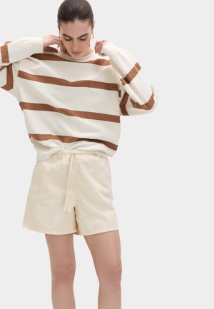 Kvinde justerer løs cream sweater med brune striber, iført lyse beige shorts med snøre, stående mod en ensartet lys baggrund.