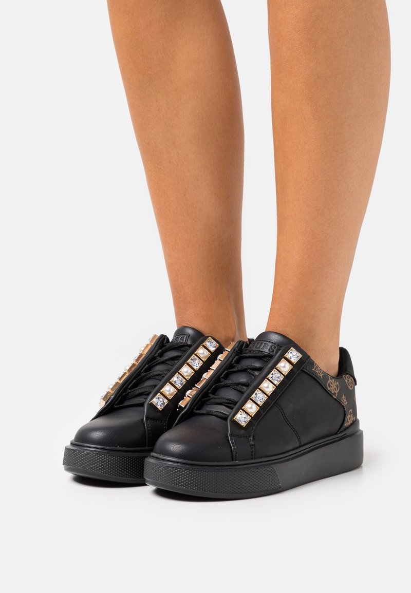 Baskets à plateforme noires avec ornements en strass et perles, portées sur des jambes nues devant un fond clair uni.