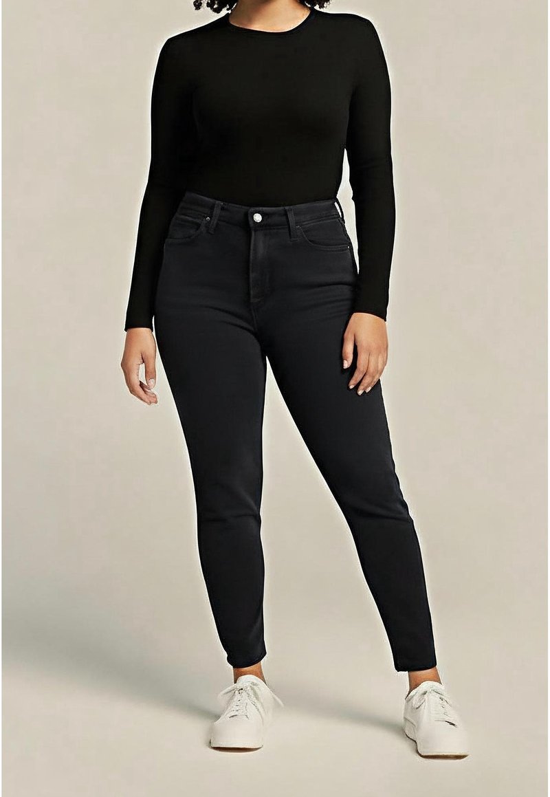 Femme portant un haut noir ajusté à manches longues, un jean noir taille haute et des baskets blanches, debout devant un fond uni.