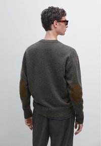 Maglione grigio a maglia con orlo e polsini a coste, caratterizzato da toppe di pelle marrone sui gomiti. Modello con occhiali da sole scuri. Design semplice e strutturato.